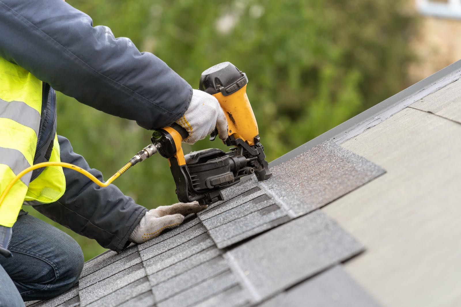 A gloved roofer's hands driving a nail into an asphalt shingle course with a pneumatic roofing nailer.
